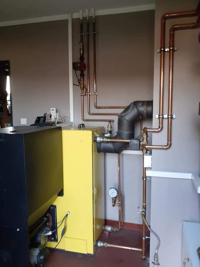 Żółto-czarny piec z podłączonymi miedzianymi rurami instalacji grzewczej, widoczne elementy pomiarowe i zawory, szare ściany.