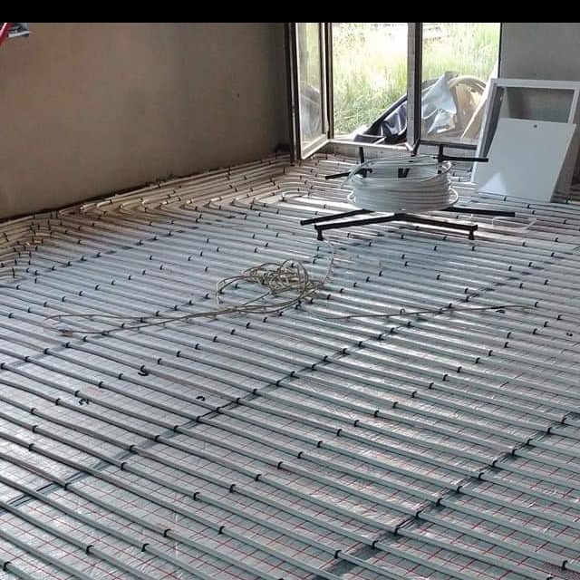 Rozłożona instalacja ogrzewania podłogowego z widoczną rolką rur i uchwytami montażowymi, przygotowana do zalania wylewką w surowym wnętrzu.