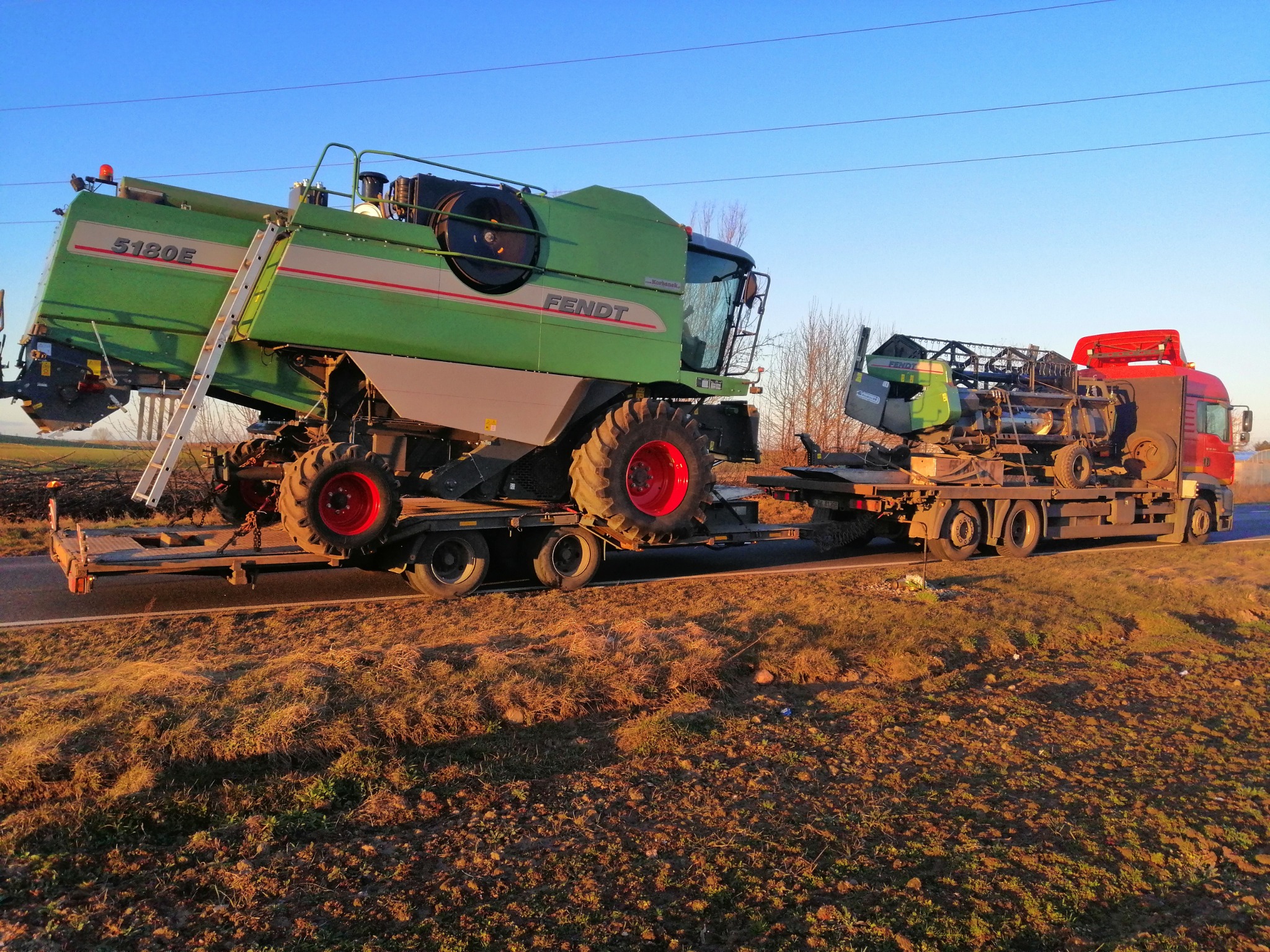 Zielony kombajn Fendt 5180E i jego heder na niskopodwoziowych naczepach ciągniętych przez czerwoną ciężarówkę, widok z boku na tle błękitnego nieba.
