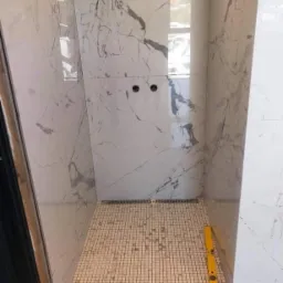 Wnętrze kabiny prysznicowej w trakcie wykańczania: białe płytki imitujące marmur na ścianach, mozaika podłogowa, żółta poziomica leżąca na podłodze, widoczne dwa otwory na baterię.