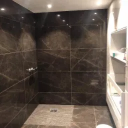 Nowoczesna kabina prysznicowa z ciemnymi, marmurkowymi płytkami, odpływ liniowy z mozaiki, oświetlenie punktowe w suficie i białe, wnękowe półki.