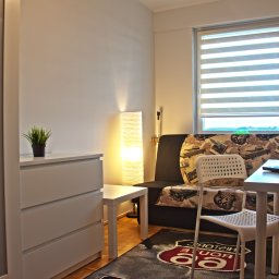 Pokojowy Najem Sp. z o.o. - Pokój w jasnych barwach z rozkładaną sofą z motywem Route 66, biurkiem z laptopem, białą komodą i lampą podłogową.