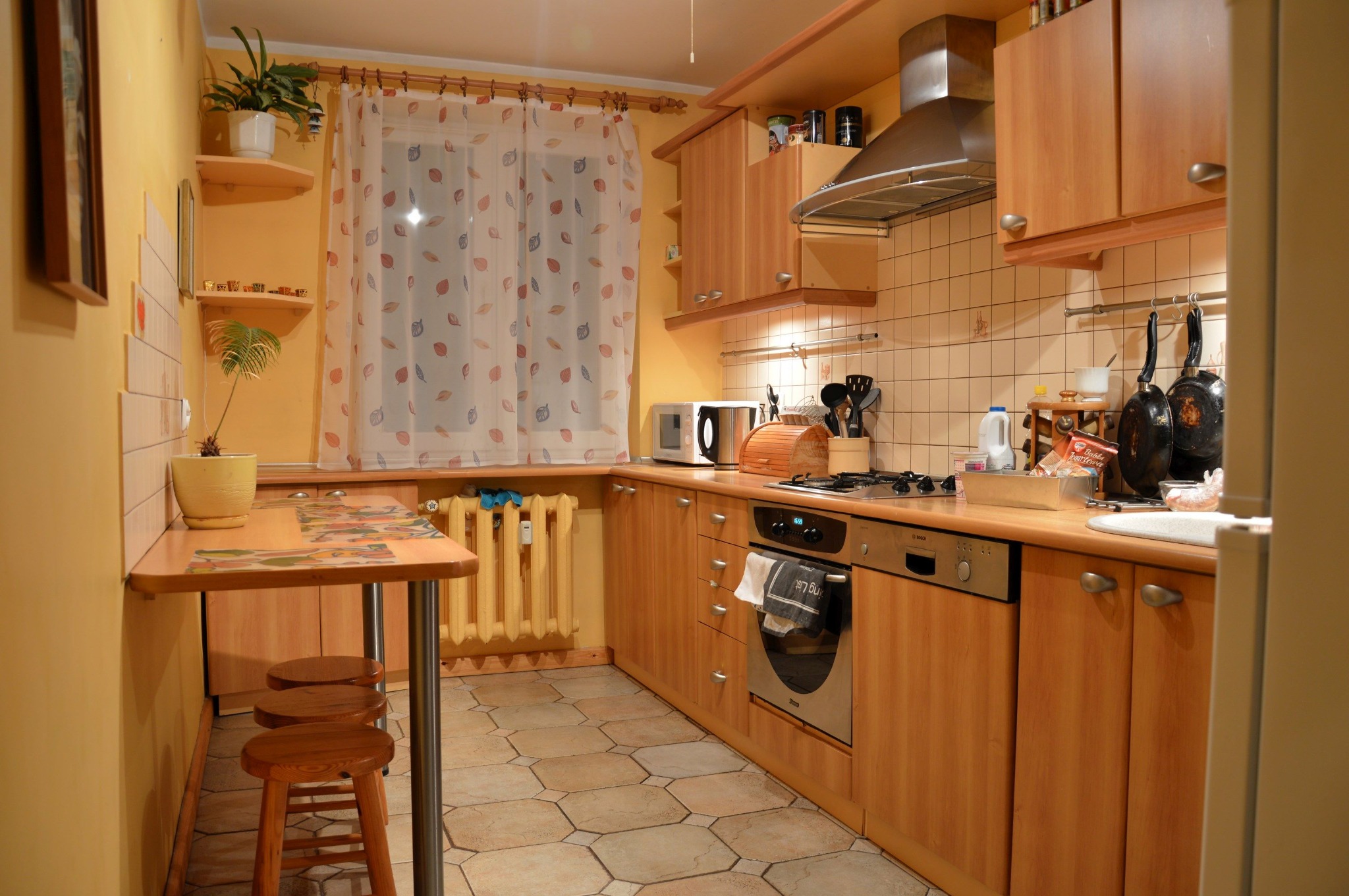 Jasna kuchnia z drewnianymi szafkami, okapem i stołem barowym z trzema stołkami, widok na okno z zasłonami w liście.