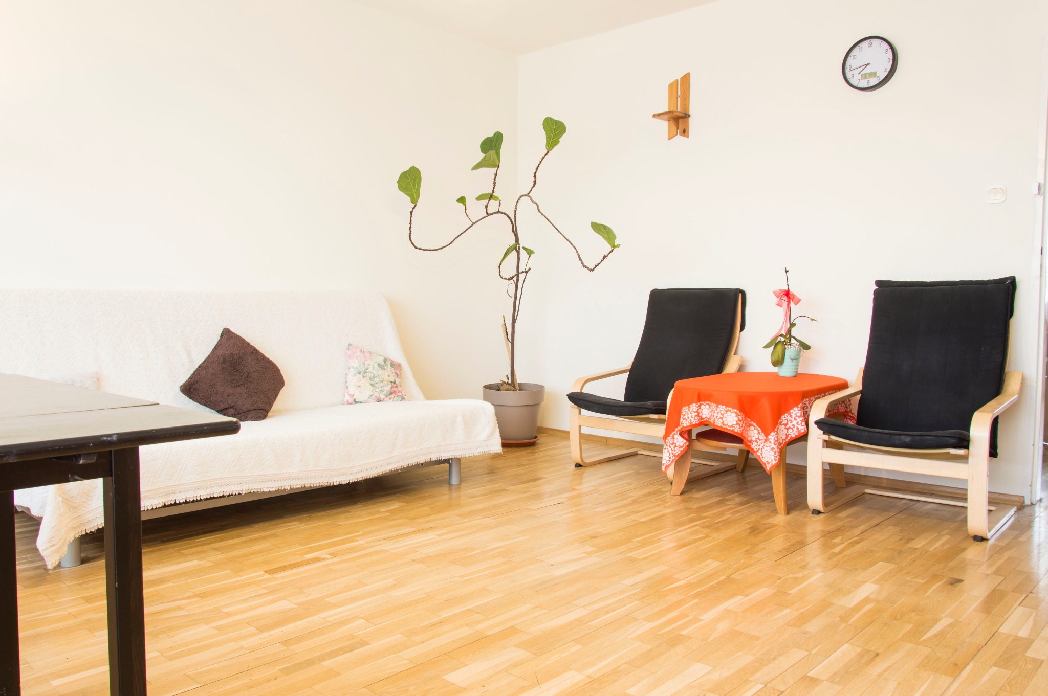 Jasne wnętrze pokoju z drewnianą podłogą, dwoma krzesłami z czarnymi poduszkami, kanapą przykrytą białą narzutą, stolikiem z pomarańczowym obrusem i rośliną w doniczce.