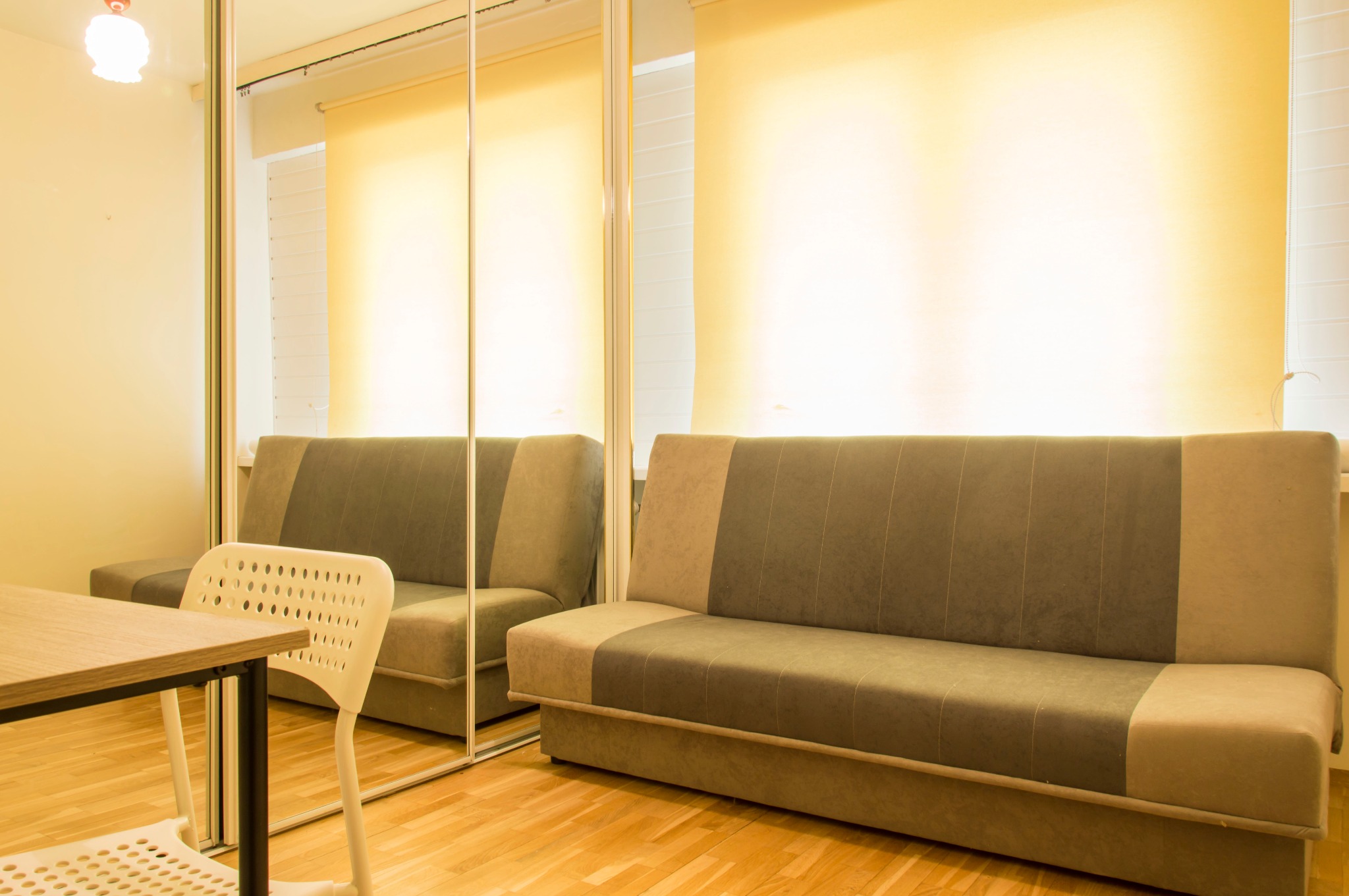 Pokój z rozkładaną sofą w odcieniach brązu i beżu, widzianą w lustrzanej szafie, z widocznym fragmentem stołu i krzesła z perforowanym oparciem.