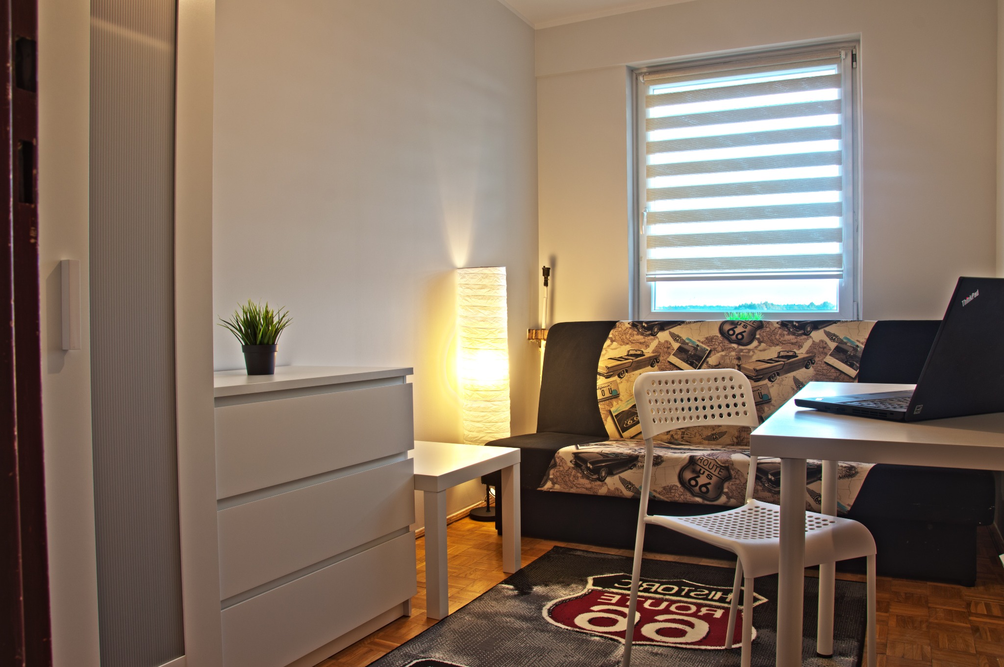 Pokój w jasnych barwach z rozkładaną sofą z motywem Route 66, biurkiem z laptopem, białą komodą i lampą podłogową.