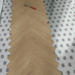 Układanie paneli podłogowych w jodełkę na podkładzie z nadrukowanymi liniami cięcia, widoczny gumowy młotek.