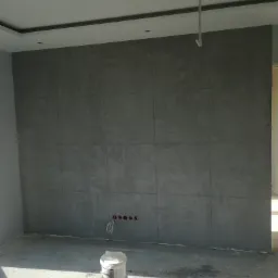 Ściana wykończona szarą, betonową fakturą, widoczne puszki elektryczne, na podłodze stoją wiadra z farbą i narzędzia malarskie, sufit podwieszany z oświetleniem punktowym.