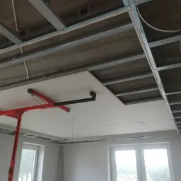 Montaż płyty gipsowo-kartonowej na stelażu sufitowym z użyciem podnośnika, widoczne kable instalacji elektrycznej.