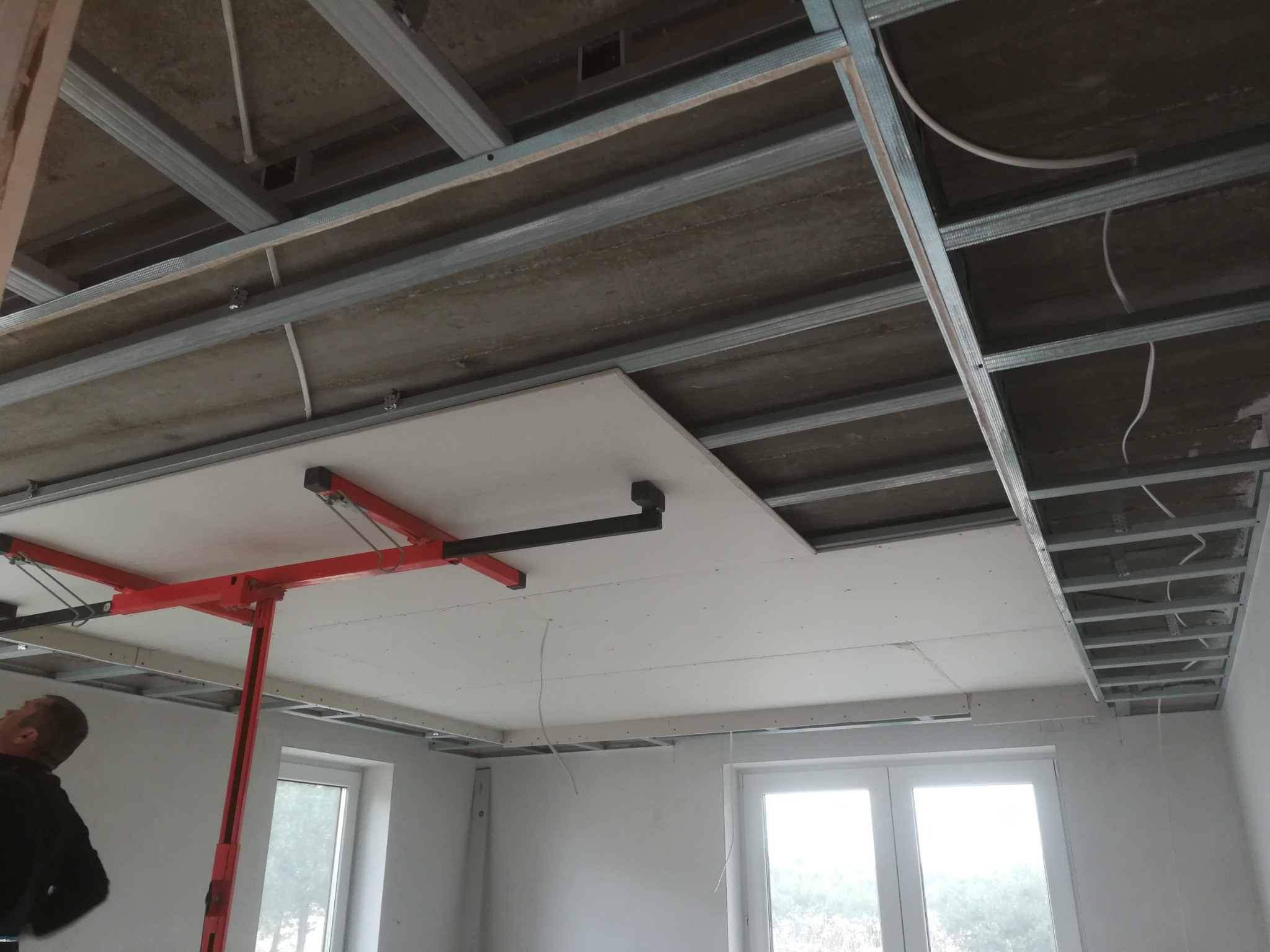 Montaż płyty gipsowo-kartonowej na stelażu sufitowym z użyciem podnośnika, widoczne kable instalacji elektrycznej.