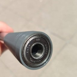 Przemyslaw Niewiadowski - Szary, cylindryczny element z łożyskiem kulkowym na końcu, widoczne oznaczenia 'FLT' i 'POLAND' na łożysku, trzymany w dłoni na tle betonowej nawierzchni.