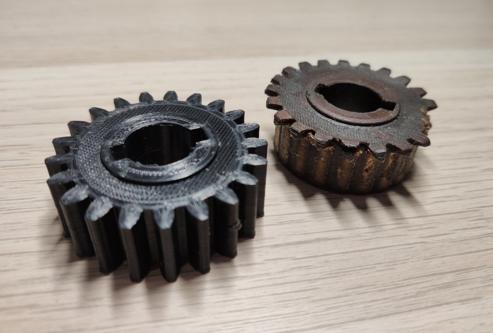 Porównanie nowej, czarnej zębatki z drukarki 3D i starej, zużytej zębatki o brązowym odcieniu, leżących na drewnianej powierzchni.