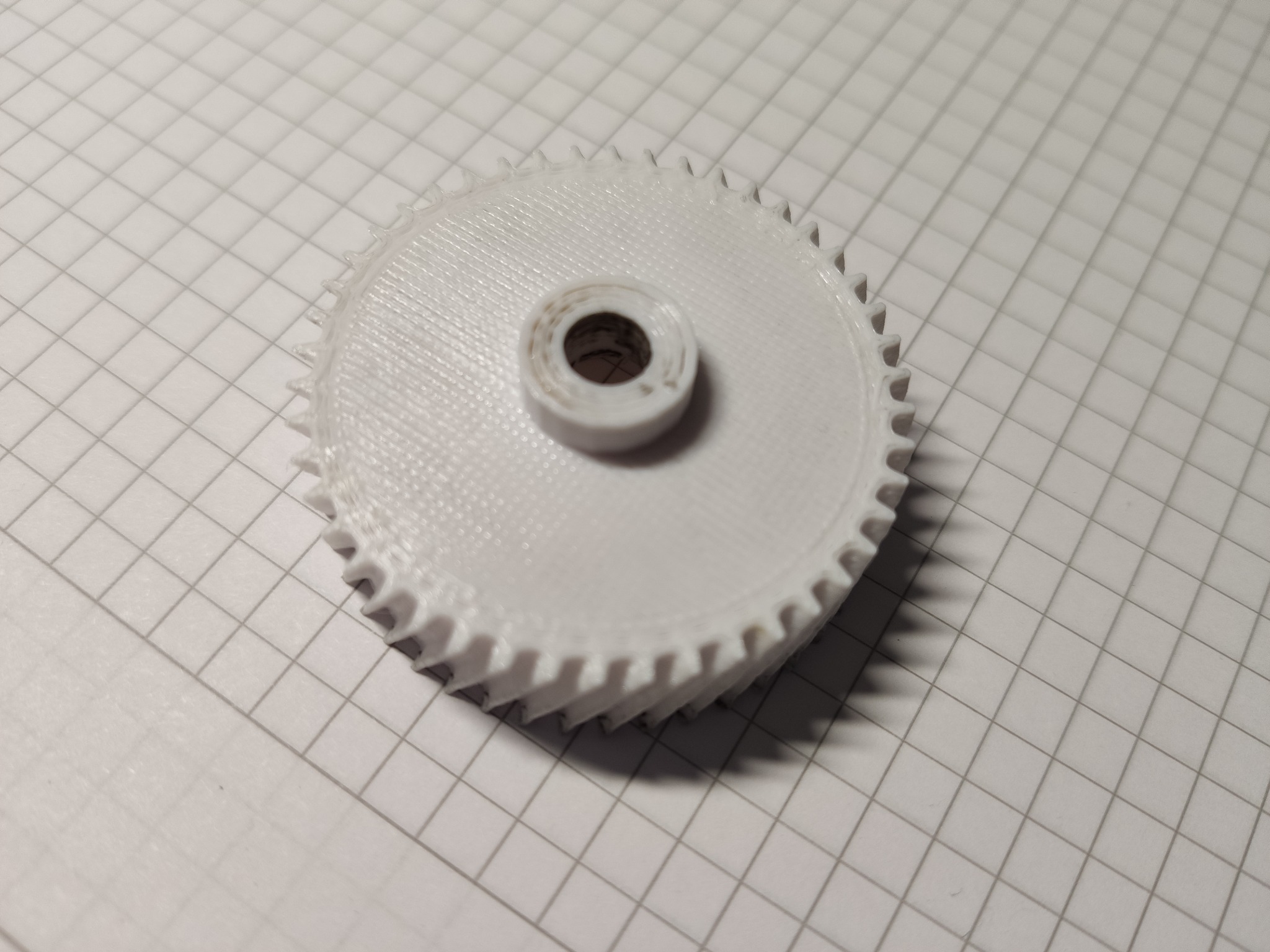 Biała, plastikowa zębatka z łożyskiem w środku, widoczne ślady druku 3D, leżąca na papierze w kratkę.