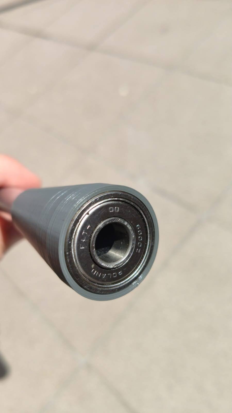 Szary, cylindryczny element z łożyskiem kulkowym na końcu, widoczne oznaczenia 'FLT' i 'POLAND' na łożysku, trzymany w dłoni na tle betonowej nawierzchni.