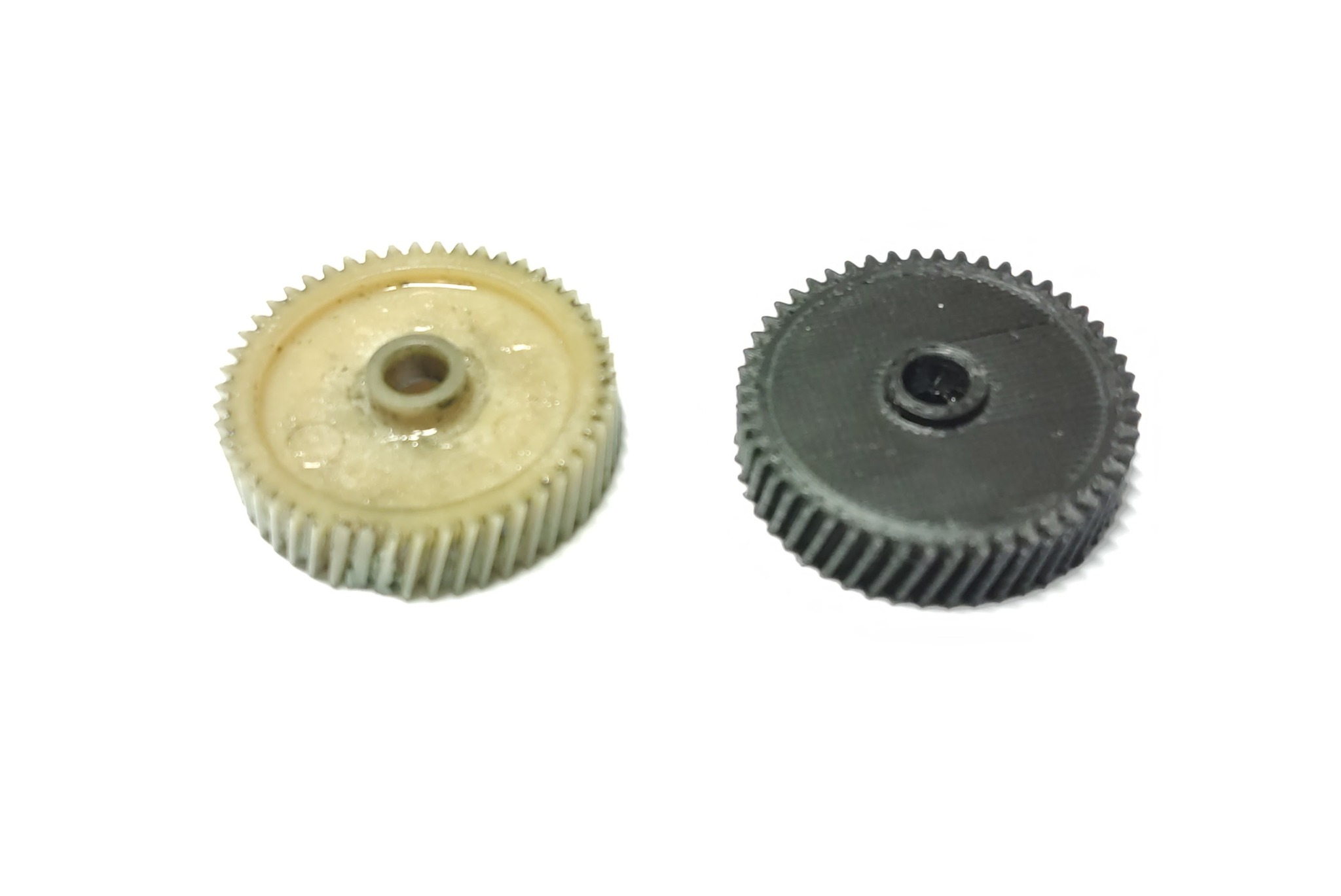 Dwie plastikowe zębatki, beżowa i czarna, leżące obok siebie na białym tle, z widocznymi śladami smaru na beżowej zębatce.