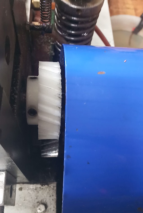 Biała, spiralna zębatka plastikowa zamontowana w mechanizmie maszyny, przylegająca do niebieskiej obudowy. Widoczne ślady smaru i drobne zabrudzenia.