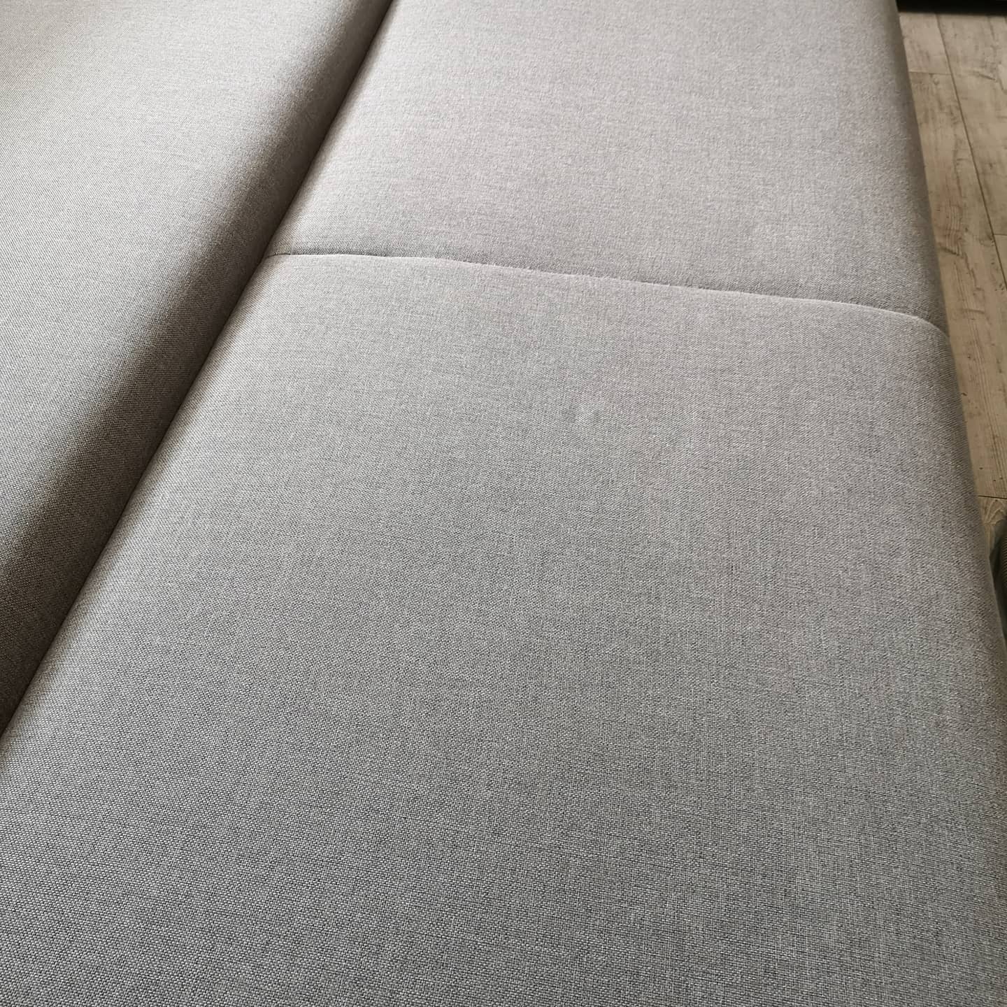 Powierzchnia jasnoszarej, tekstylnej tapicerki sofy z widocznym szwem łączącym elementy siedziska, lekko zarysowana faktura materiału.