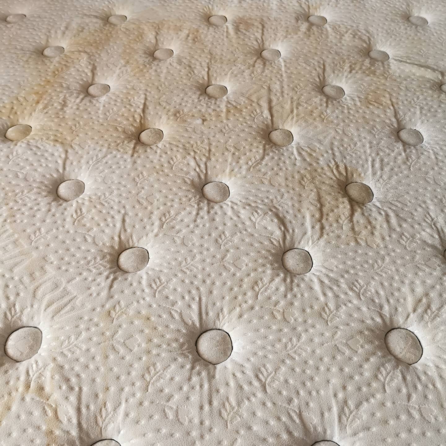 Materac z jasną tapicerką, widoczne plamy i zabrudzenia, wzór w liście, regularne pikowania z guzikami.