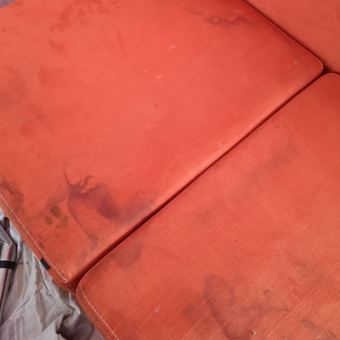 Pomarańczowe poduszki kanapy z widocznymi, ciemnymi plamami i zabrudzeniami, wymagające odświeżenia. Częściowo widoczne elementy konstrukcyjne kanapy i poszarzała tkanina w tle.