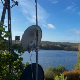 Dwie anteny satelitarne zamontowane na słupie z widokiem na jezioro i jesienny las w tle, z bliska widoczny maszt energetyczny i zieleń drzew.