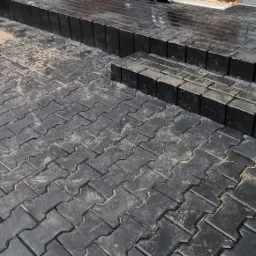 Ułożona, czarna kostka brukowa typu 'Jodełka' tworząca schody wejściowe, z widocznymi śladami piasku na powierzchni i mokrymi stopniami.