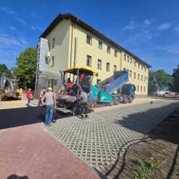 Układanie asfaltu na parkingu z ażurowych płyt betonowych. W tle budynek, ekipa robotników i maszyny budowlane: walec i ciężarówka z wywrotką.