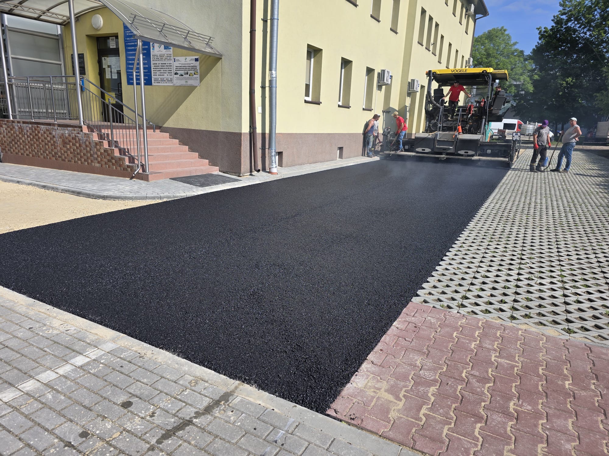Świeżo położony asfalt na parkingu przy budynku użyteczności publicznej. Widoczna maszyna do asfaltowania i pracownicy. Część parkingu wyłożona jest ażurową kostką.