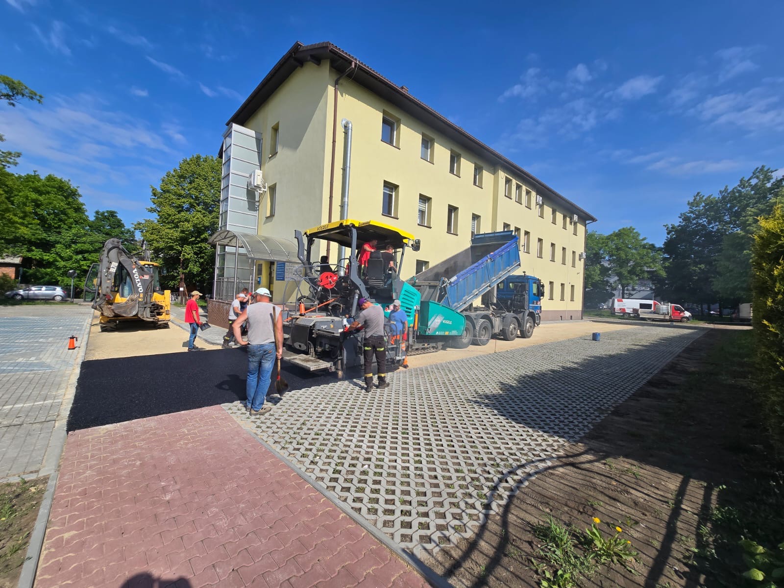 Układanie asfaltu na parkingu z ażurowych płyt betonowych. W tle budynek, ekipa robotników i maszyny budowlane: walec i ciężarówka z wywrotką.