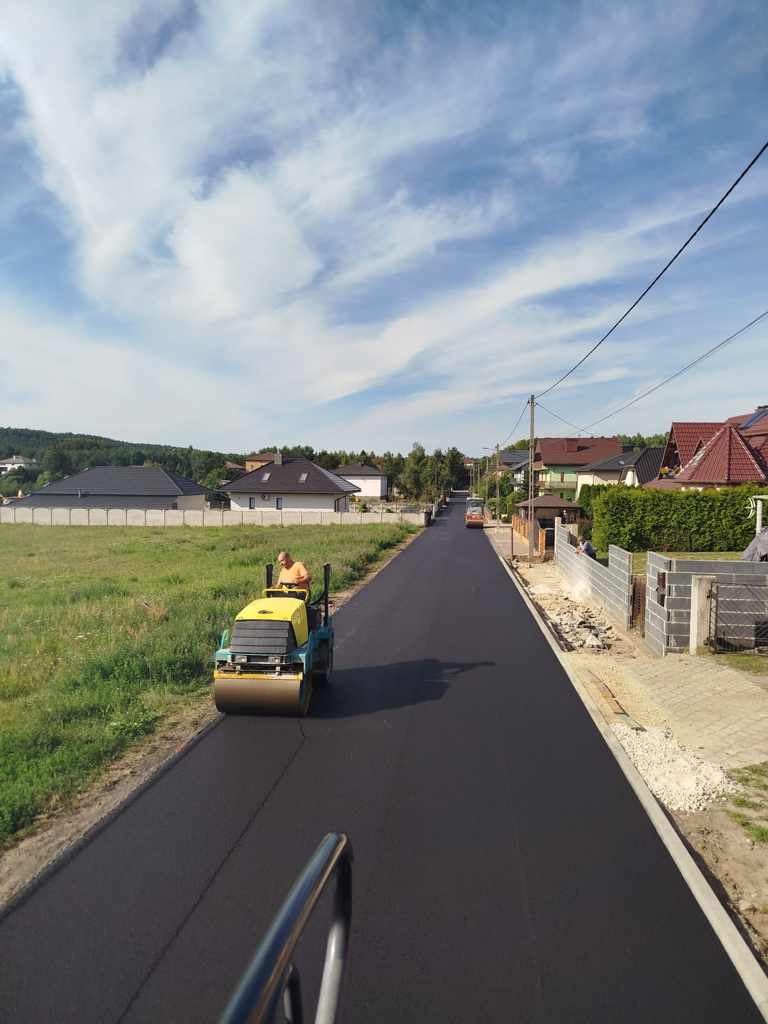 Walec drogowy ubija świeżo wylany asfalt na ulicy w zabudowie jednorodzinnej, błękitne niebo z chmurami w tle, widok z perspektywy operatora walca.