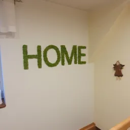 Zielony napis HOME z mchu na białej ścianie nad schodami z drewnianą barierką, widoczne oświetlenie sufitowe i mała dekoracja anioła.
