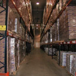 Wnętrze magazynu z wysokimi regałami wypełnionymi paletami z zapakowanymi kartonami, zabezpieczonymi folią stretch, widok w perspektywie wzdłuż korytarza.