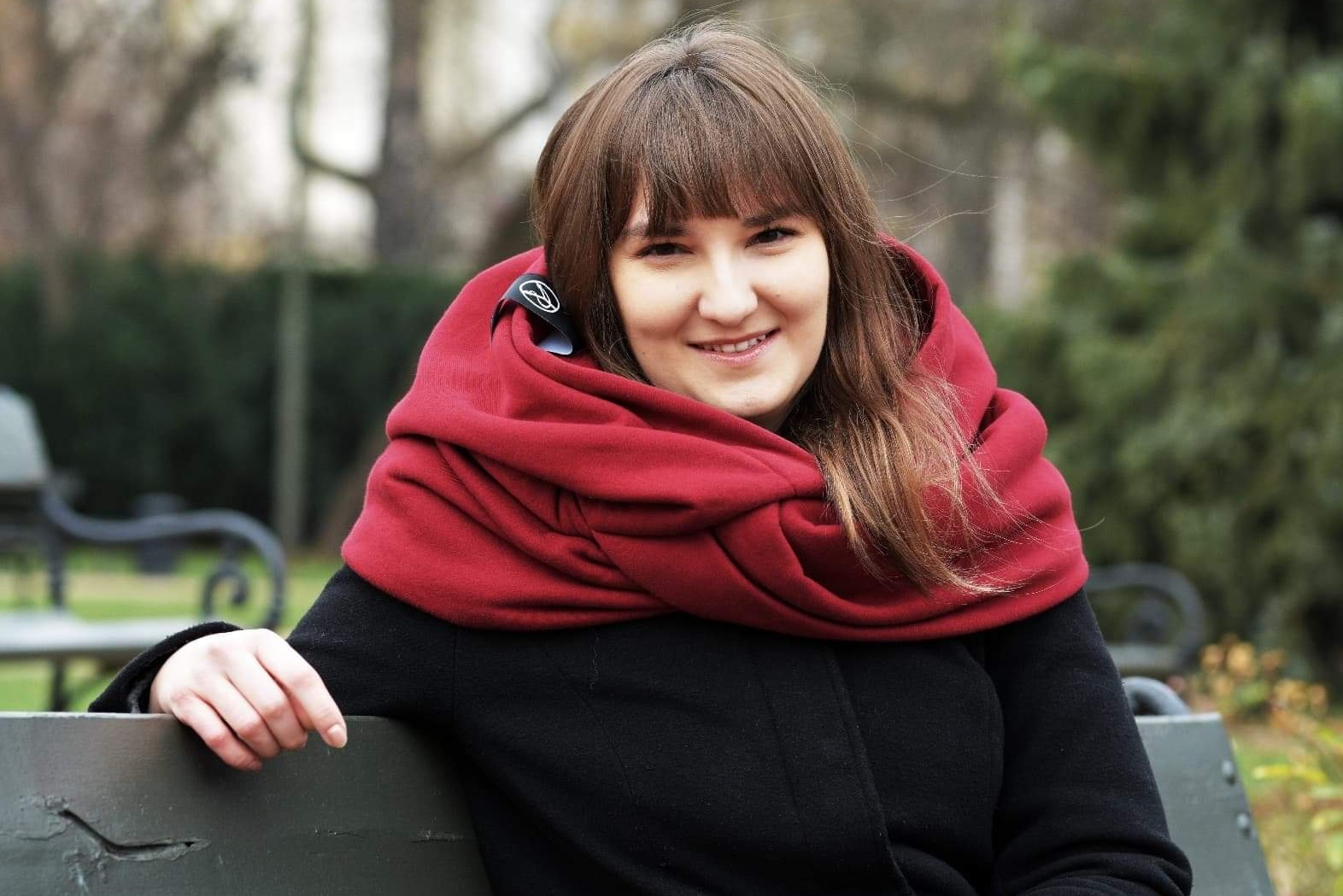 Uśmiechnięta kobieta w czarnym płaszczu i bordowym, grubym szalu z widocznym logo producenta na klipsie, siedząca na ławce w parku.