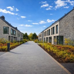 BDKD Sp. z o.o. - Osiedle domów z szarej cegły z brukowaną drogą i zielenią. Widok w perspektywie, błękitne niebo z chmurami w tle. Prestiżowa inwestycja.