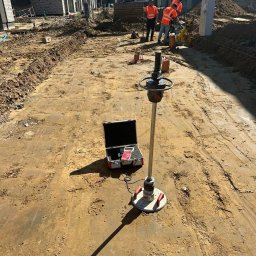 BDKD Sp. z o.o. - Prace przygotowawcze pod budowę drogi: ubijanie gruntu specjalistycznym sprzętem pomiarowym, w tle ekipa budowlana i koparka na placu budowy osiedla.