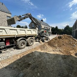 BDKD Sp. z o.o. - Ciężarówka z wywrotką i koparka na placu budowy, w tle nowoczesne domy z cegły. Widoczny etap przygotowania terenu pod budowę drogi lub układanie kostki.