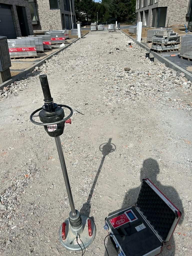 Etap budowy drogi: ubijak dynamiczny i otwarty kufer z urządzeniem pomiarowym na podsypce z kruszywa, w tle palety z kostką brukową i budynki.