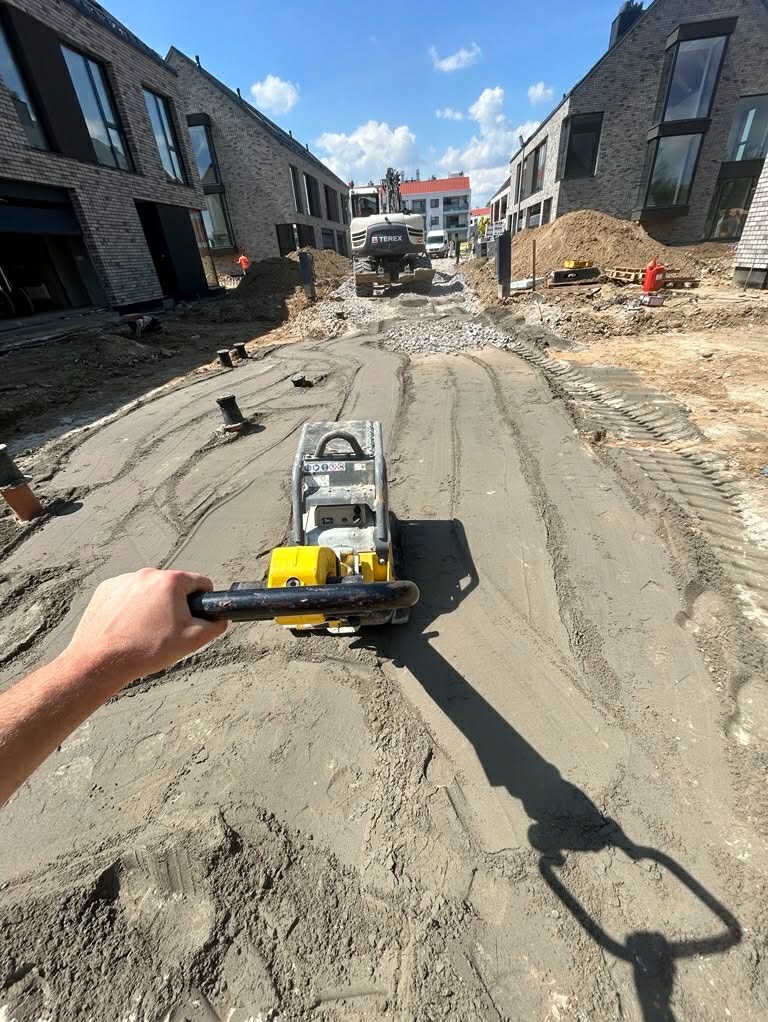 Praca zagęszczarką na placu budowy drogi osiedlowej. W tle budynki z cegły i maszyny budowlane. Widok z perspektywy operatora zagęszczarki.