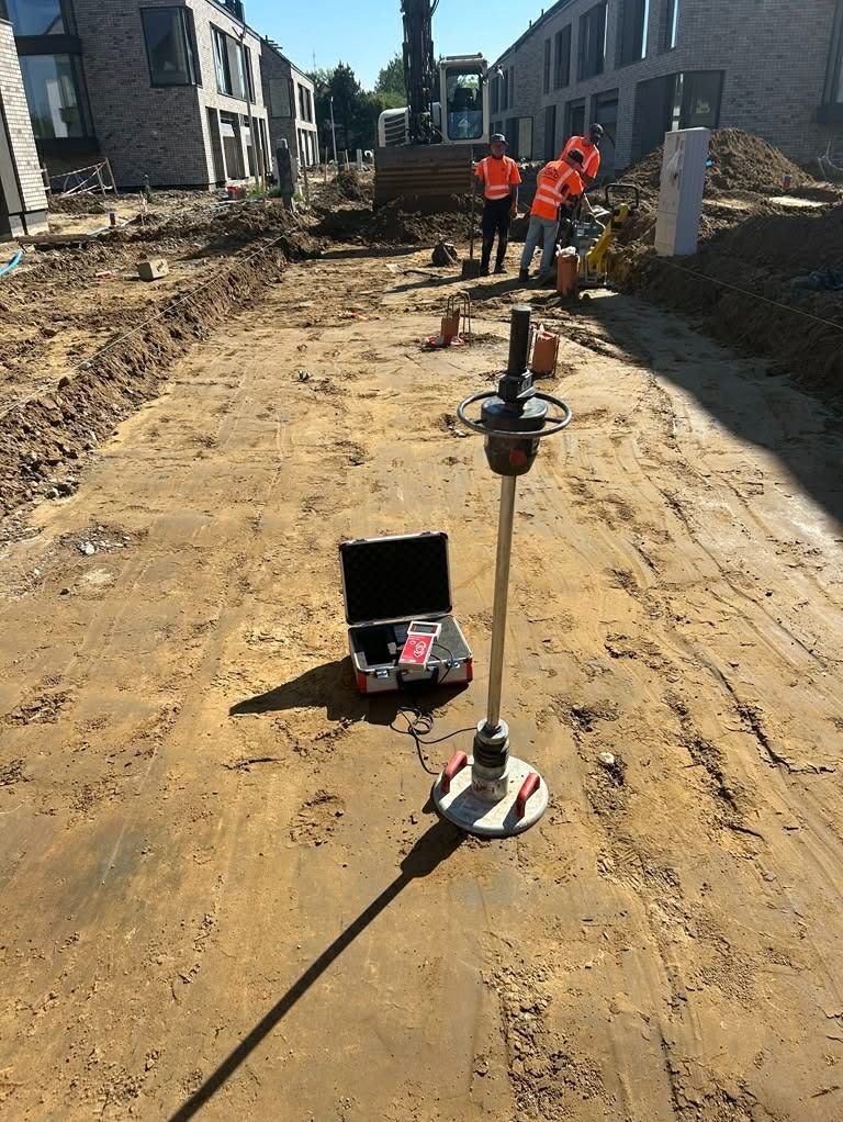 Prace przygotowawcze pod budowę drogi: ubijanie gruntu specjalistycznym sprzętem pomiarowym, w tle ekipa budowlana i koparka na placu budowy osiedla.