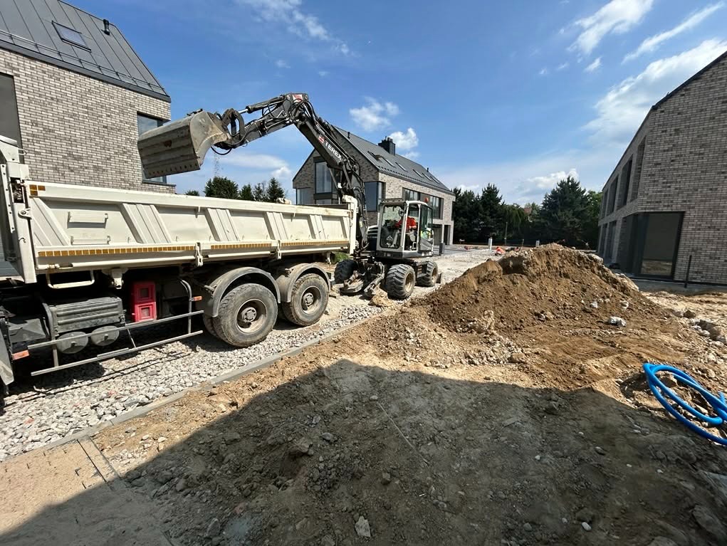 Ciężarówka z wywrotką i koparka na placu budowy, w tle nowoczesne domy z cegły. Widoczny etap przygotowania terenu pod budowę drogi lub układanie kostki.