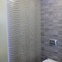 Wykończona łazienka z toaletą, ściany wyłożone szarymi płytkami o falującej strukturze i prostokątnymi płytkami, oświetlenie LED wzdłuż krawędzi ściany.