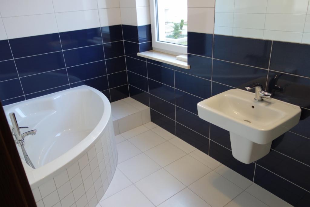 Wykończona łazienka w bieli i granacie, z asymetryczną wanną, umywalką na postumencie i oknem z widokiem na zieleń.