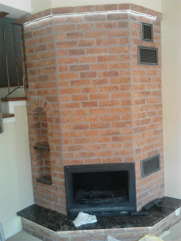 Kanciasty kominek z cegły z czarnym marmurowym blatem, oświetleniem LED i kratkami wentylacyjnymi, widok z dołu.