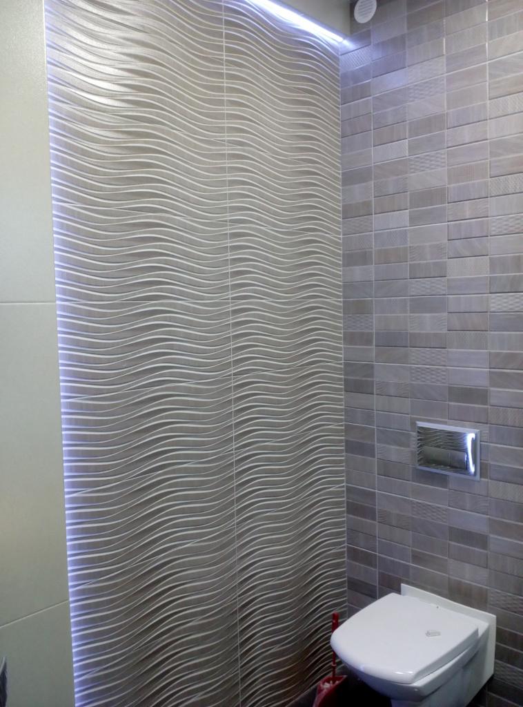 Wykończona łazienka z toaletą, ściany wyłożone szarymi płytkami o falującej strukturze i prostokątnymi płytkami, oświetlenie LED wzdłuż krawędzi ściany.