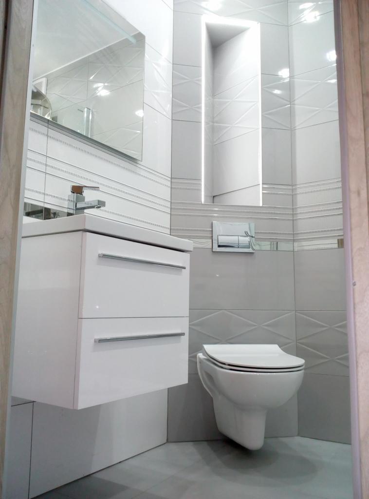 Nowoczesna, jasna łazienka z podwieszaną toaletą, białą szafką pod umywalką z chromowanymi uchwytami i lustrem z podświetleniem, wykończona szarymi i białymi płytkami z geometrycznym wzorem.