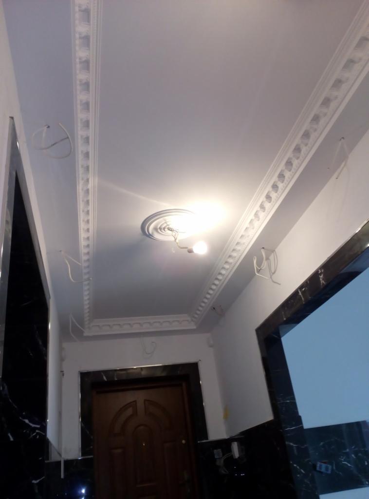 Wykończony sufit z dekoracyjnymi listwami przysufitowymi i centralnym elementem dekoracyjnym wokół punktu oświetleniowego, widoczne wystające przewody elektryczne przed ostatecznym montażem lampy.
