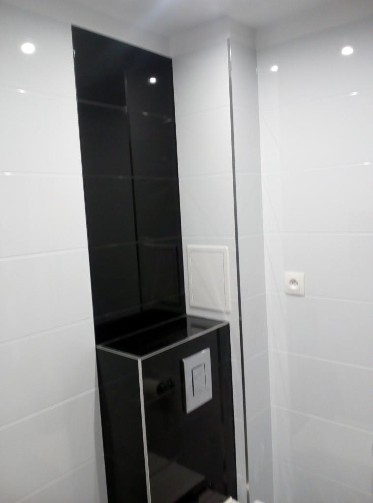 Nowoczesna łazienka z czarno-białymi płytkami, zabudową toalety z czarnego szkła i białymi ścianami, widoczny fragment instalacji elektrycznej.