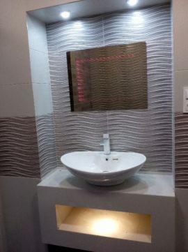 Umywalka nablatowa o owalnym kształcie, osadzona na białej szafce z wnęką oświetloną od dołu, na tle ściany wyłożonej płytkami imitującymi falę, z lustrem w ramie, oświetlona punktowo.