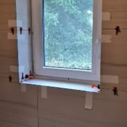 Montaż płytek wokół okna w łazience, widoczne poziomice i krzyżyki dystansowe, płytki imitujące drewno ułożone poziomo.