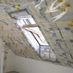 Montaż folii paroizolacyjnej Isover Vario KM Duplex UV na poddaszu, widoczny fragment okna dachowego i białe ściany.