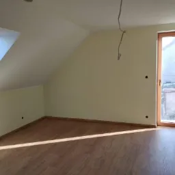 Poddasze po remoncie: świeżo pomalowane ściany w odcieniu kremowym, drewnopodobna podłoga, okno dachowe i drzwi balkonowe z widokiem na zewnątrz, wystające kable ze ściany.